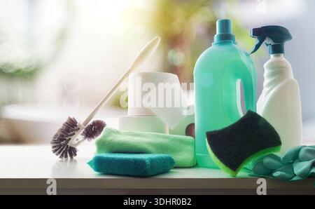 Bathroom cleaning supplies such as disinfectant bottles, scouring pads, gloves, toilet brush and toilet paper on white wooden bench and bathroom in th Stock Photo