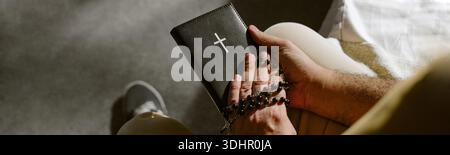 Header of Man Praying Holding Bible and Rosary Beads Indoors Stock Photo