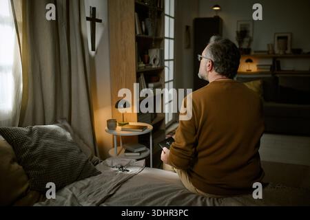 Middle Aged Man Praying Alone While Sitting on Bed at Home Stock Photo