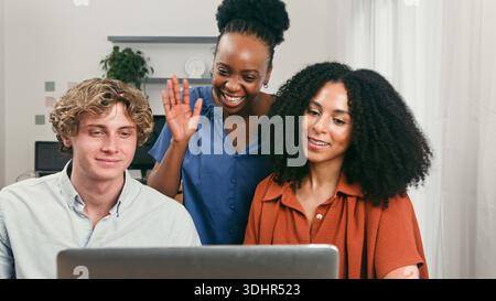 Three Diverse Professionals Collaborate On Laptop, Smiling And Waving In A Startup Office Stock Photo