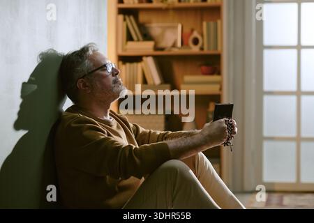Middle Aged Man Sitting Against Wall Holding Rosary and Praying Stock Photo