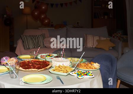 Table displaying party food including pizza, cake, chips, candy and donuts with empty chairs in living room setting, suggesting preparation for celebration or gathering Stock Photo