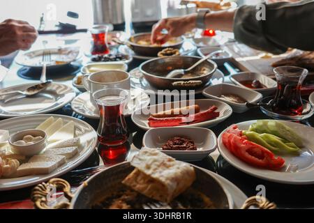 Rich traditional Turkish breakfast spread with cheeses, olives, sausages, tomatoes, cucumbers, pastries, jams and black tea in tulip glasses, hands se Stock Photo