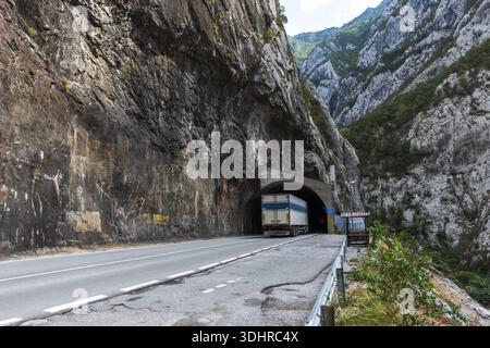 A large truck nears a dark tunnel carved through towering rock. The winding road hugs a rugged cliffside with greenery and distant peaks, blending ind Stock Photo