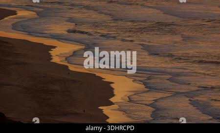 View of golden light reflecting on the sandy beach and ocean waves, with silhouettes of people strolling along the shore, Wilderness, Western Cape, So Stock Photo