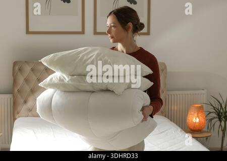 Woman holding pile of soft white folded duvet and pillows at home in her bedroom, cozy domestic lifestyle, housewife cleaning, tidying up bedroom, hou Stock Photo