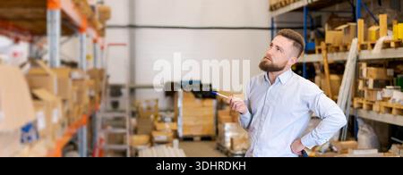 A bearded warehouse manager stands with hand on hip, pen in hand, thoughtfully looking upward at shelves. Captures strategic thinking, inventory asses Stock Photo