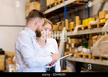 Man and woman logistics managers working together in a warehouse. Bearded man writing on a clipboard while colleague looks on. Concept of teamwork, st Stock Photo