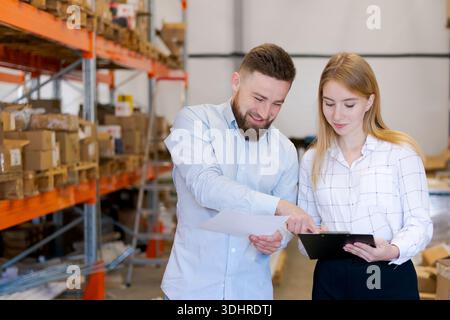 Smiling man and woman logistics supervisors discussing paperwork and pointing at a clipboard in a warehouse. Concept of successful teamwork, distribut Stock Photo
