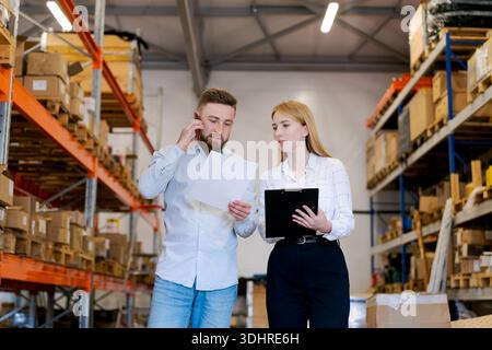 Professional man talking on mobile phone while female colleague reviews documents on a clipboard in a warehouse. Low angle shot showing industrial sto Stock Photo