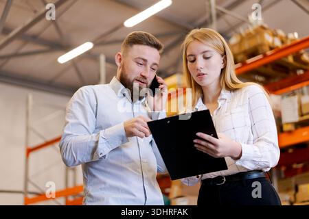 Professional logistics team in a distribution center. Man talks on a smartphone while woman reviews data on a clipboard. Concept of industrial managem Stock Photo