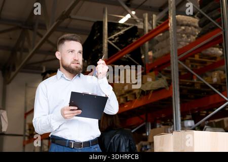 Male logistics supervisor holding a clipboard and pen while looking up thoughtfully. Professional manager overseeing stock control and distribution st Stock Photo