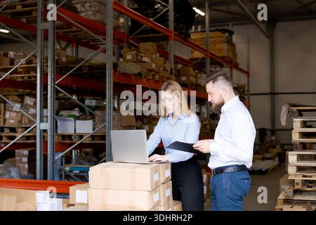 Male and female logistics managers collaborating in a distribution center. Woman uses a laptop on cardboard boxes while man reviews data on a clipboar Stock Photo