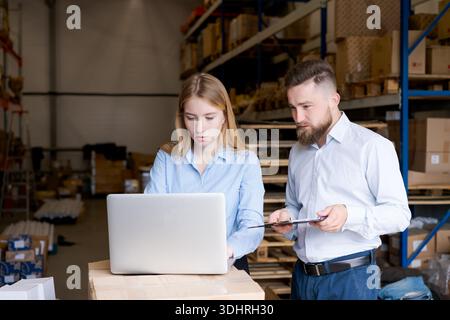 Focused man and woman supervisors using a laptop on a parcel box to track inventory. Professional team coordinating supply chain operations in a distr Stock Photo