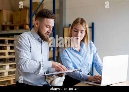 Male supervisor and female manager discussing data while using a laptop and clipboard in a warehouse. Professional team coordinating logistics, shippi Stock Photo