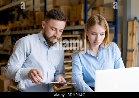 Two warehouse employees working together on inventory management. Man and woman checking stock data using laptop and clipboard in modern storage facil Stock Photo