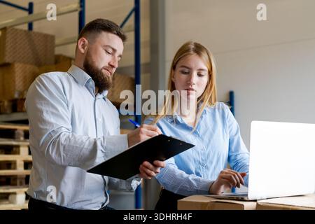 Male and female logistics supervisors collaborating in a warehouse. They are checking stock data using a laptop and writing notes on a clipboard. Conc Stock Photo
