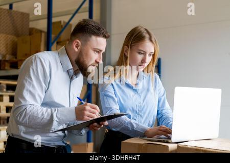 A professional team of logistics managers, a man and a woman, working together in a warehouse. They are checking inventory records using a laptop and Stock Photo
