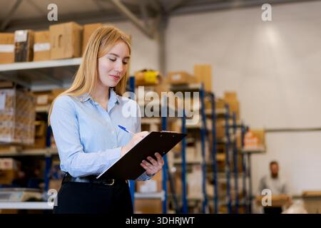 Professional female logistics supervisor writing notes on a clipboard in a large warehouse. Stock audit and inventory control process with a worker an Stock Photo