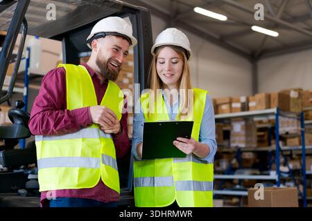 Smiling warehouse managers in safety gear reviewing documents on a clipboard. Logistics team in hard hats and reflective vests standing near a forklif Stock Photo