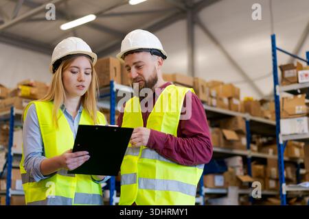 Female manager and male worker in safety helmets and reflective vests checking stock data on a clipboard. Professional logistics team coordinating ope Stock Photo
