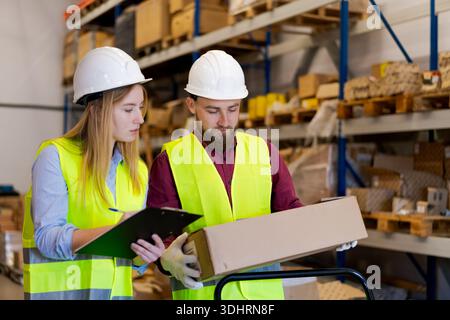 Focused male and female workers examining package details while recording inventory data on clipboard. Meticulous quality control and verification pro Stock Photo
