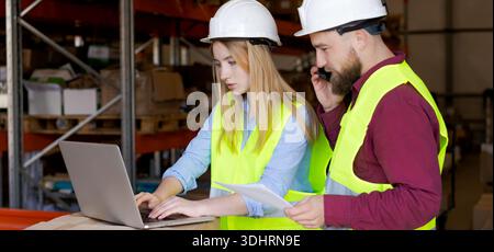 Focused female employee working on computer while male coworker handles client call. Digital workflow efficiency and multitasking in warehouse operati Stock Photo