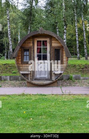 A whimsical circular wooden vacation cabin sits among birch trees in a green park. The rustic roundhouse features a weathered door, small windows, and Stock Photo