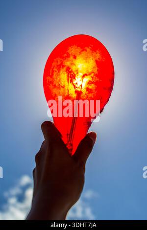 Hand holding red light bulb against clear blue sky, shot from below Stock Photo