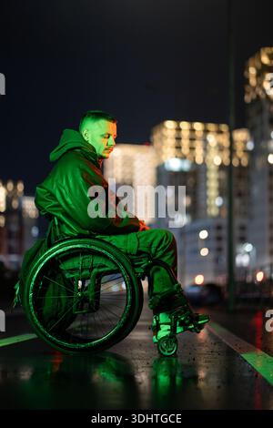 Lonely man with disability in wheelchair in rgb neon color light on late night walk outside. Stock Photo