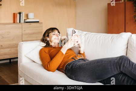 Young woman smiling, watching content on smartphone while lying on a couch at home Stock Photo
