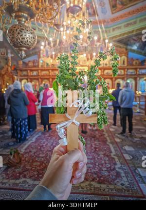 Christian hand holding a wooden cross adorned with fresh basil and white ribbon, symbol for the Exaltation of the Holy Cross. Scene inside an Orthodox Stock Photo