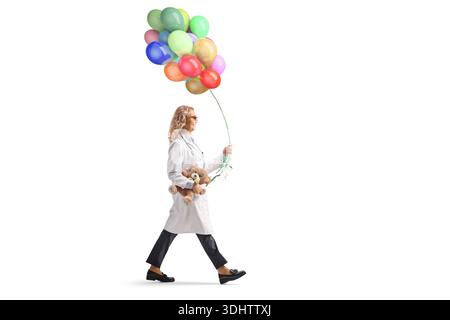 Pediatric doctor is walking with a teddy bear and holding a bunch of balloons isolated on white background Stock Photo