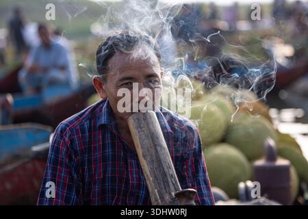 Rangamati, Bangladesh - 19 June 2023: View of a man exhaling a stream of smoke from a large wooden pipe, his face etched with concentration against a Stock Photo