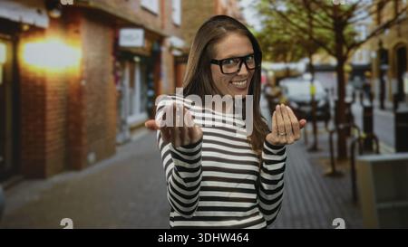 Woman with glasses beckons with both hands on a busy city street, smiling broadly; welcoming playfulness casual. Stock Photo
