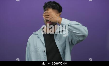 Young hispanic man wearing denim shirt covers eyes with raised hand in studio against purple background; introspection. Stock Photo