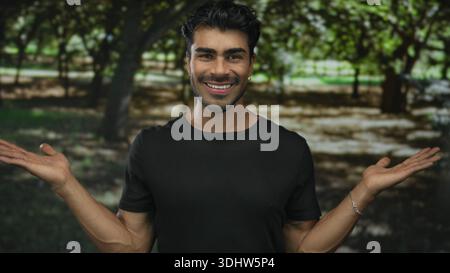 Man shows open palms with raised forearms in sunlit green forest park among tall oak trees; happiness. Stock Photo