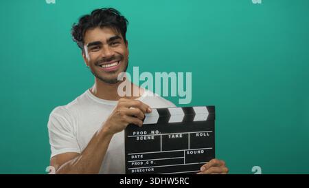 Hispanic man in white t shirt holding film clapboard against vibrant green studio wall with confident posture; creativity imagination inspiration inno Stock Photo