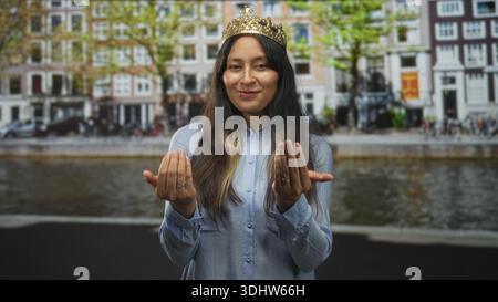 Hispanic woman wearing a gold crown smiles and beckons with both hands on amsterdam street; playful charm. Stock Photo