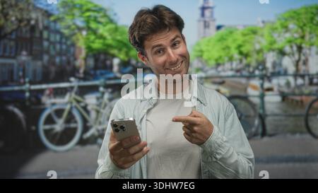 Man smiles and points finger to smartphone while holding it near canal bicycles on amsterdam street; digital connection joy. Stock Photo