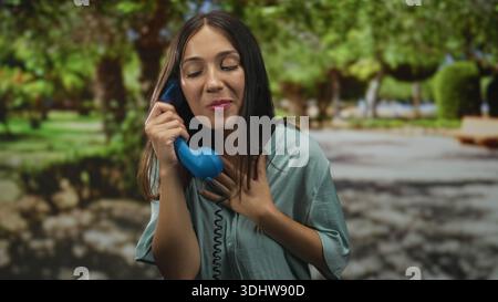 Woman holds blue telephone receiver to ear while hand rests over chest in park; gratitude connection. Stock Photo