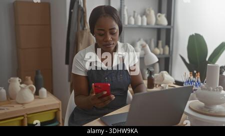 Woman potter in studio holds red phone and works at laptop, hands checking messages on pottery table; concentration craft business. Stock Photo
