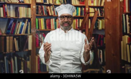 Man chef, middle aged hispanic with grey hair, wearing chef uniform and hat, holding whisk and gesturing with hands in building library; playful surpr Stock Photo