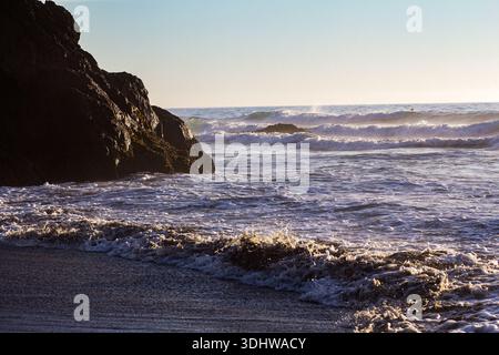 View of sunlight catching the crests of waves rolling onto a dark sandy beach, contrasting with the rugged, shadowed rocks, Fort Bragg, California, Un Stock Photo