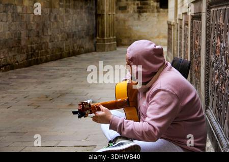 A young man in a mauve hoodie plays an acoustic guitar while sitting in a historic stone alleyway with traditional carved wooden doors. Stock Photo