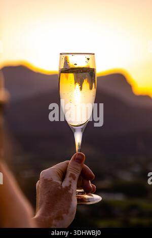 Man holding a champagne flute with white wine against a golden sunset and mountain background. Celebration or romantic evening concept. Stock Photo