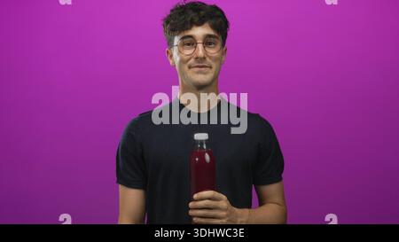 Man young hispanic holding a bottle with hand and cheeks puffed in purple studio wearing glasses and black shirt; playful curiosity. Stock Photo