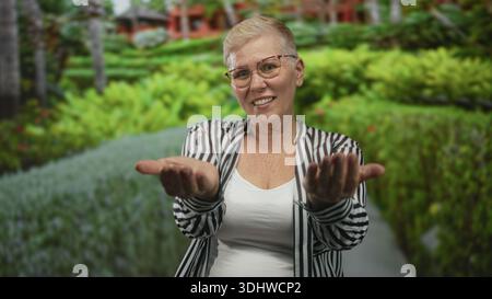 Woman beckons with both hands and visible palms on a forest path near green foliage, smiling at camera in striped shirt and glasses; welcoming invitat Stock Photo