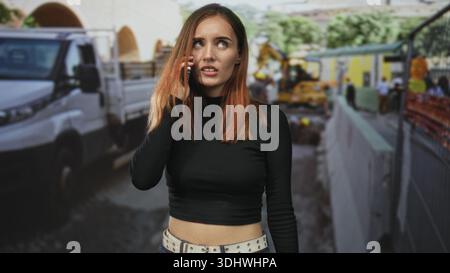 Young redhead woman in black top holds phone to ear and gestures with hand in a busy street construction zone by a white truck and barricade; frustrat Stock Photo