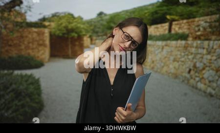 Woman holding clipboard and wearing glasses, hand on neck showing discomfort by the stone entrance of a building outdoors; work fatigue. Stock Photo